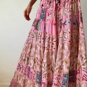 May include: A long pink skirt with a patchwork design featuring floral and geometric patterns. The skirt has a drawstring waist and a tiered silhouette.