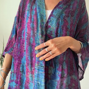 Könnte beinhalten: Ein leichter Kimono mit offener Vorderseite und einem lebendigen, abstrakten Muster in tiefem Violett, Magenta und leuchtendem Blau. Der Stoff hat eine subtil schimmernde Textur mit komplexen, dunklen, wirbelnden Liniendetails im Design.