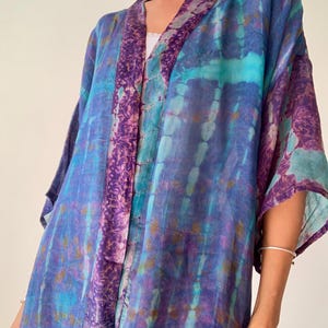 Può includere: Un kimono tie-dye fluido nei toni del blu, turchese e viola. Il kimono presenta un bordo fantasia lungo il bavero e le maniche. Il tessuto appare leggero e arioso, suggerendo un capo confortevole.