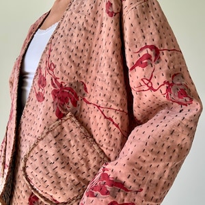 May include: A pale pink jacket featuring a red and brown floral design. The jacket has a pocket and is detailed with small, evenly spaced black stitches. The jacket is open at the front.