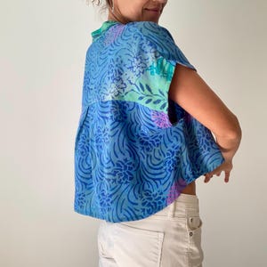 May include: A blue patterned top with short sleeves. The top features a floral and swirl design in shades of blue, green, and purple. The model is wearing white pants.