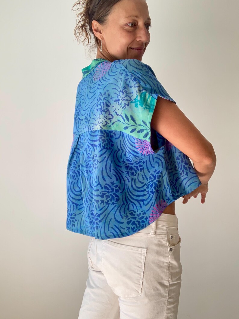 May include: A blue patterned top with short sleeves. The top features a floral and swirl design in shades of blue, green, and purple. The model is wearing white pants.