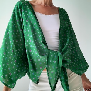Peut inclure: Un haut de style kimono vert avec un motif floral bleu, violet et blanc. Le haut est noué à la taille et a des manches larges. Le tissu semble léger et fluide.