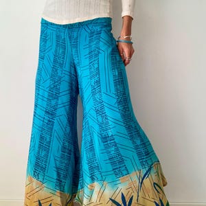 Könnte beinhalten: Weite Hose in leuchtendem Türkis mit geometrischem Muster. Der untere Saum hat einen beigen Rand mit dunkelblauen Blattmotiven. Die Hose wird mit einem cremefarbenen Langarm-Oberteil getragen.