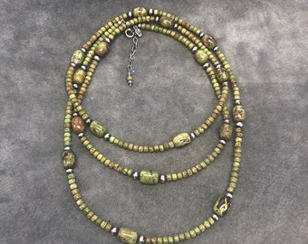 May include: A multi-strand necklace made of green and silver beads. The necklace features a variety of green beads, some with a speckled pattern, and silver beads that are interspersed throughout the necklace. The necklace is layered, with three strands of beads.
