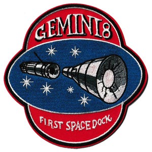 May include: Embroidered patch with the Gemini 8 mission logo. The design features a red border with the text "GEMINI 8" and "FIRST SPACE DOCK". A blue oval in the centre depicts a spacecraft docking, surrounded by white stars.