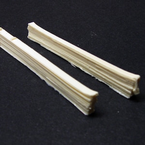 May include: Two long, thin, off-white, wooden strips, each with a layered, stacked, and slightly uneven texture. The strips are lying flat on a black surface.