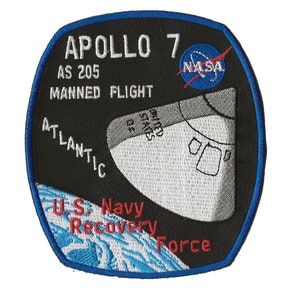 May include: A black and blue embroidered patch with the text "APOLLO 7 AS 205 MANNED FLIGHT" and a NASA logo. The patch also features a white illustration of a spacecraft with the text "UNITED STATES OF" and "ATLANTIC U.S. Navy Recovery Force".