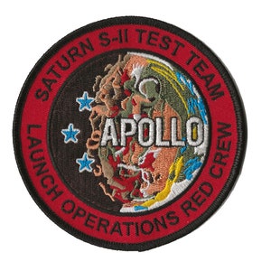 Pode incluir: Um remendo bordado vermelho, branco e azul com o texto "Saturn S-II Test Team Launch Operations Red Crew" que envolve um gráfico de lançamento de foguetes com a palavra "Apollo" em texto branco.