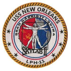 May include: A white embroidered patch with a gold rope border. The patch features a white circle with the text "USS NEW ORLEANS" in black. Inside the circle is a red, white, and blue image of a man with outstretched arms. The text "BEAN GARRIOTT LOUGIMA" is written around the image. Below the image is the text "SKYLAB II" in red, white, and blue. The text "LPH-11" is written below the image.