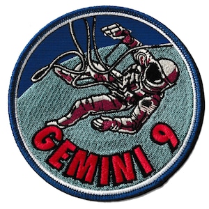 Op de afbeelding: Een cirkelvormige geborduurde patch met een astronaut in een bordeauxrood ruimtepak die in de ruimte zweeft. De patch heeft een blauwe achtergrond met het woord "GEMINI" in rood en het nummer "9". De patch is omrand met een blauw-witte gestikte rand.