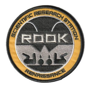 May include: A circular embroidered patch featuring a black and gold border. The center displays the text ROOK in silver, with the words SCIENTIFIC RESEARCH STATION arched above and RENAISSANCE below in black. A stylized silver ant graphic is visible.