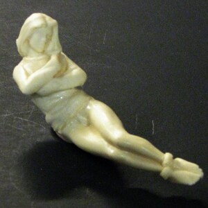 May include: A white ceramic figurine of a woman lying on her side with her arms crossed in front of her.