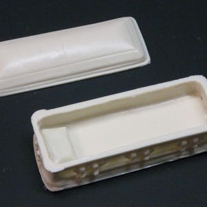May include: A white plastic coffin with a lid. The coffin has a decorative design around the edges.