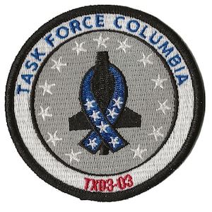 May include: A grey and white circular patch with the text "Task Force Columbia" in a white circle around a black silhouette of a plane with a blue ribbon with white stars. The text "TX03-03" is in red at the bottom of the patch.