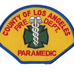 Può includere: Una toppa ricamata gialla e blu con il testo "County of Los Angeles Fire Dept. Paramedic" e una croce verde con un serpente bianco avvolto attorno.