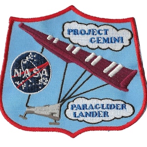 Op de afbeelding: Een blauwe en rode geborduurde patch met een wit NASA-logo, een rode en witte zweefvlieger en de tekst "PROJECT GEMINI" en "PARAGLIDER LANDER".