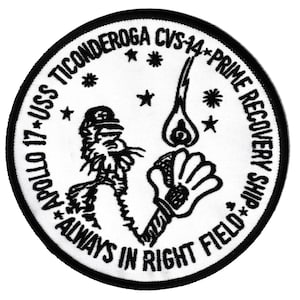 May include: A circular patch with a black border and white interior. The patch features text including "Apollo 17", "USS Ticonderoga", and "Prime Recovery Ship". A cartoon figure and a hand holding a flame are also depicted.