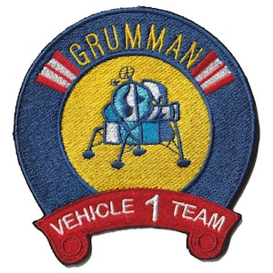 Op de afbeelding: Geborduurde patch met een blauw en geel cirkelvormig ontwerp, het woord "GRUMMAN" bovenaan. Het midden toont een blauw-witte maanlander. Een rode banner onderaan leest "VEHICLE 1 TEAM".