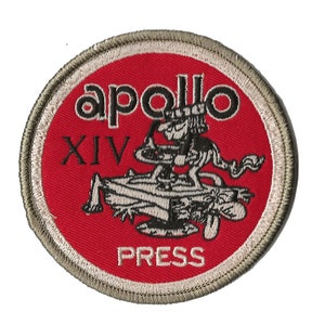May include: A red and white embroidered patch with the text "apollo XIV PRESS" and a cartoon image of a man in a hat sitting on a rocket.