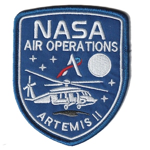 May include: A blue and white embroidered patch with the NASA logo and "AIR OPERATIONS". The patch shows a helicopter, stars, and the text "ARTEMIS II". The patch is shield-shaped.