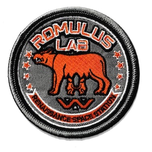 以下が含まれることがあります： オレンジと白で「ROMULUS LAB」の文字が入った丸いパッチ。中央にはオレンジ色のクマのような生き物がいます。「RENAISSANCE SPACE STATION」の文字が下部にあります。パッチには黒い縁取りがあります。