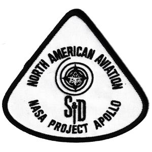 May include: A white, triangular patch with black embroidered text and a black border. The patch reads "NORTH AMERICAN AVIATION" and "NASA PROJECT APOLLO" around a central emblem. The emblem features a target design with a stylized image.