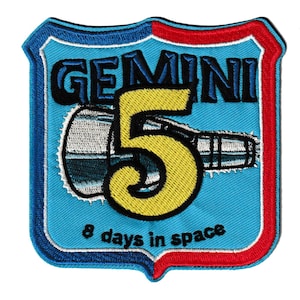 May include: Embroidered patch with the Gemini 5 mission logo. The shield-shaped patch has a light blue background and a red and blue border. The number "5" is yellow, and the text "GEMINI" and "8 days in space" are displayed.