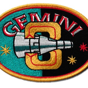 Op de afbeelding: Geborduurde patch met het woord "GEMINI" in rood en wit, en een gestileerd ruimteschipontwerp. De ovale patch heeft een turquoise en zwarte achtergrond, een gouden rand en steraccenten.