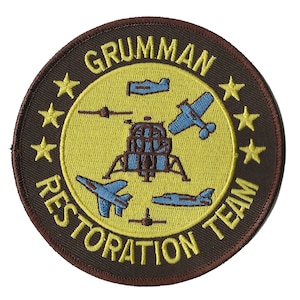 May include: A brown circular patch with a yellow circle in the centre. The yellow circle has the text "GRUMMAN RESTORATION TEAM" in a yellow outline around the edge. Inside the yellow circle are five blue aeroplanes and a brown lunar module.