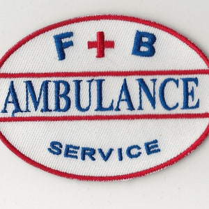Puede incluir: Parche bordado de forma ovalada con fondo blanco. El parche presenta las palabras "AMBULANCE SERVICE" en azul, con "F + B" arriba y una cruz roja. Rayas rojas y blancas bordean el parche.