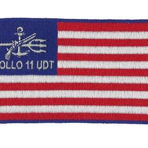 May include: Embroidered patch featuring a design of the American flag with red and white stripes and a blue field. The blue field includes a trident, anchor, and text that reads "APOLLO 11 UDT". The patch has a blue border.