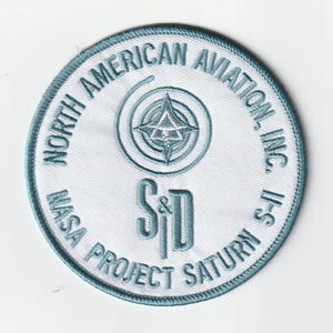 May include: A circular embroidered patch with a white background and blue border. The text reads NORTH AMERICAN AVIATION, INC. and NASA PROJECT SATURN S-II. In the center is a stylized star emblem above the large blue letters S&D.