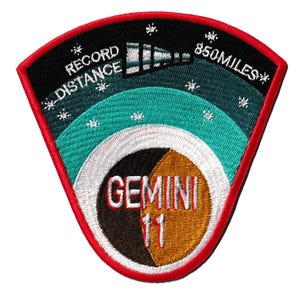 May include: Embroidered patch with a red border, featuring the text "RECORD DISTANCE" and "850 MILES". The design includes a telescope, stars, and a segmented circle with "GEMINI 11" in the center, with black, teal, and gold colors.