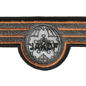 Op de afbeelding: Een geborduurde patch in grijs en oranje met een gestileerde bloem en de tekst "JAKDF" in een cirkel. De patch heeft drie oranje strepen op een grijze achtergrond.