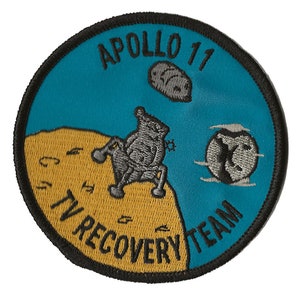 May include: A round, blue and yellow embroidered patch with the text "APOLLO 11 TV RECOVERY TEAM" and a cartoon image of a lunar module landing on the moon.