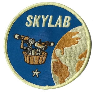 May include: A blue and white embroidered patch with the word "SKYLAB" in white lettering. The patch features a cartoon image of two astronauts in a space capsule orbiting a brown and white Earth.