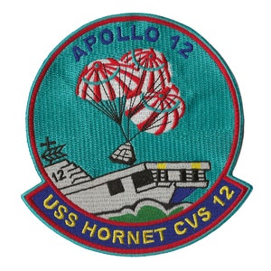 May include: A blue and white embroidered patch with the text "Apollo 12 USS Hornet CVS 12" and a depiction of a spacecraft with parachutes descending onto an aircraft carrier.