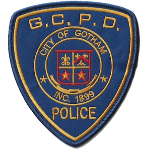 May include: A shield-shaped embroidered patch in blue with gold borders. It features the text G.C.P.D. at the top, CITY OF GOTHAM in a circle, INC. 1899, and POLICE at the bottom. A central crest displays red stars and gold symbols on a blue field.