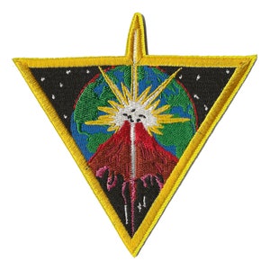 Op de afbeelding: Een gele driehoekige patch met een zwarte omtrek en een witte ster in het midden. De ster wordt omringd door een rode, groene en blauwe wereld met een zwarte achtergrond. De wereld wordt omringd door een ring van witte sterren.