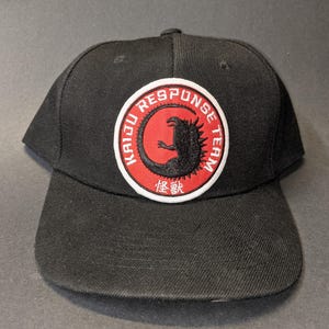 Könnte beinhalten: Schwarze Baseballkappe mit einem roten und weißen gestickten Aufnäher, auf dem "Kaiju Response Team" steht, mit einer schwarzen Silhouette eines Monsters im Kreis. Die japanischen Schriftzeichen "怪獣" befinden sich unterhalb des Kreises.