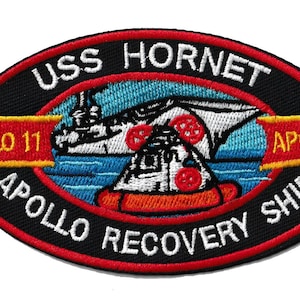 May include: Embroidered patch featuring the USS Hornet, Apollo 11, and Apollo 12. The patch has a black oval border with red and yellow accents. The design includes a depiction of a spacecraft and the text "APOLLO RECOVERY SHIP."