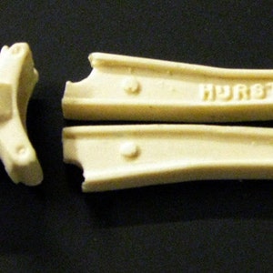 May include: A set of off-white plastic model car parts, including a rear axle housing, two drive shafts, and two suspension arms. The suspension arms are labelled "HURST".