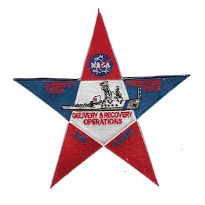 May include: A red, white and blue star-shaped patch with the text "Liberty Star" and "Freedom Star" on the top points. The centre of the star features a ship with the text "Delivery & Recovery Operations" and the NASA logo. The bottom points of the star have the text "KSC" and "CCAFS".