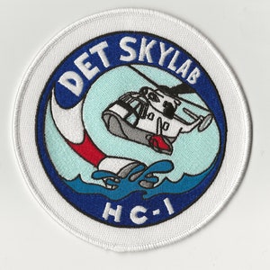 May include: A white embroidered patch with a blue circle containing a white helicopter flying over blue water with the text "DET SKYLAB HC-I" in white.
