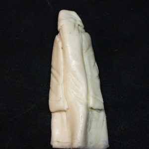 May include: A white ceramic figurine of a person wearing a long robe and a hat. The figurine is standing upright and has a simple design.