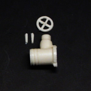 Op de afbeelding: Een witte plastic modelkit van een klep met een wiel, twee kleine pijpen en een grotere pijp. Het model is niet gemonteerd.