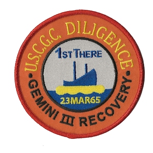 May include: A round, orange patch with a red border. The patch has the text "USCG.C. DILIGENCE" in yellow around the edge. Inside the circle is a white background with a blue ship and the text "1ST THERE" above it. The text "23MAR65" is below the ship. The text "GEMINI III RECOVERY" is in black around the bottom of the circle.