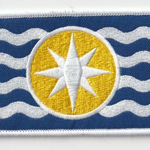 May include: Embroidered patch featuring a blue background with white wavy lines and a central yellow circle. Inside the circle is a white eight-pointed star. The patch has a white border.