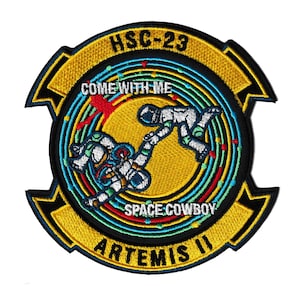 May include: Embroidered patch with a circular design featuring two astronauts, one reaching out to the other. The patch includes the text "HSC-23", "COME WITH ME", "SPACE COWBOY", and "ARTEMIS II" in various colors, set against a yellow and blue background.